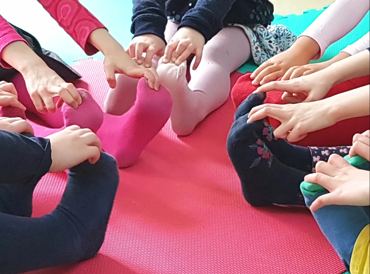 Kinder sitzen im Kreis auf einer Yogamatte und fassen sich an die Füße